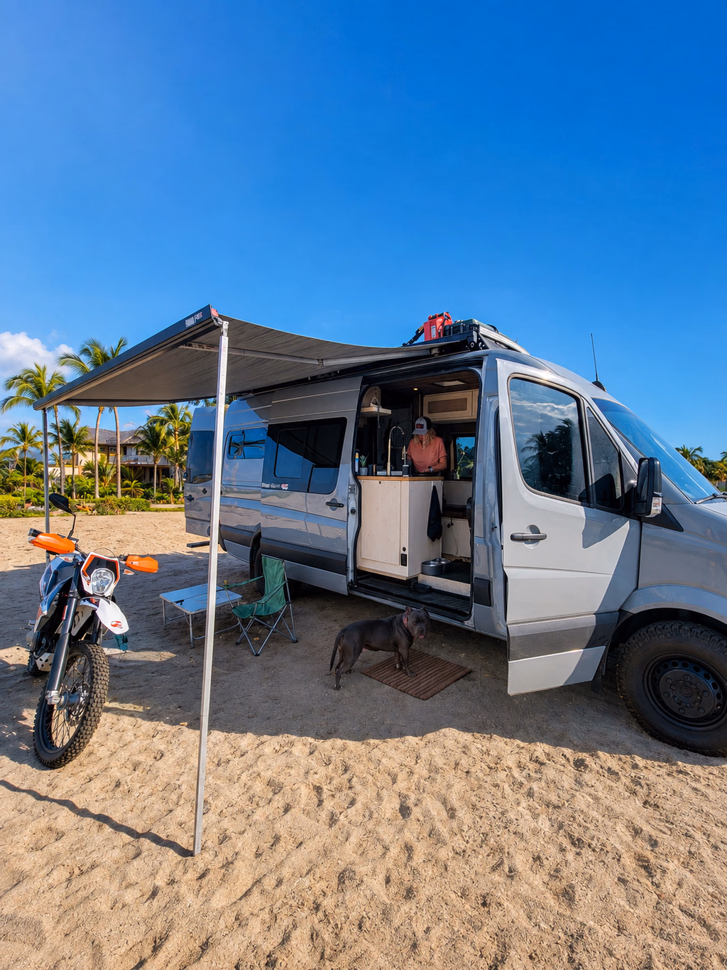 Fourgon aménagé installé sur une plage avec l’auvent déployé et la moto prête à partir.